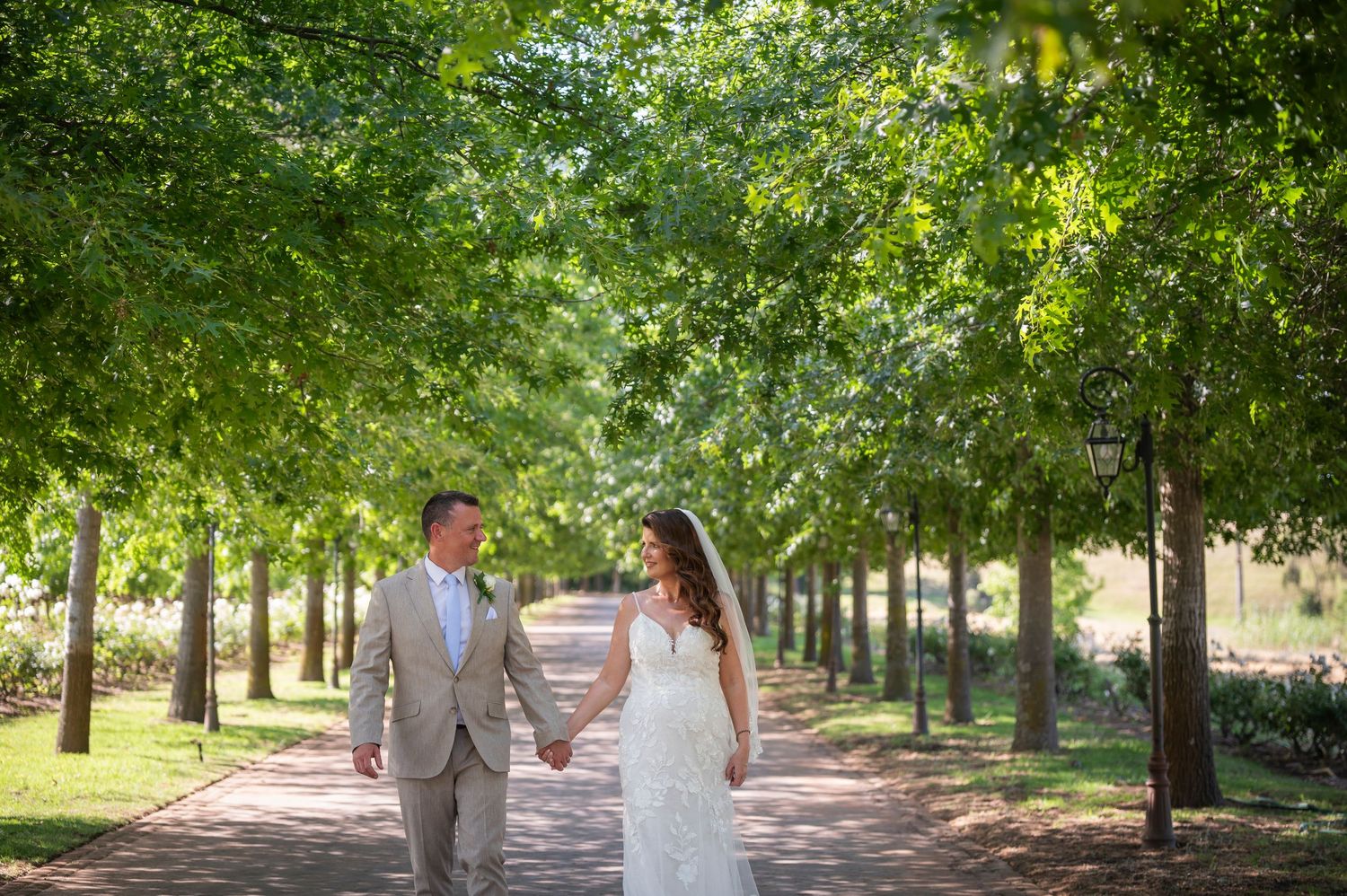Bride and groom walk hand-in-hand down a tree-lined brick path on their wedding day.