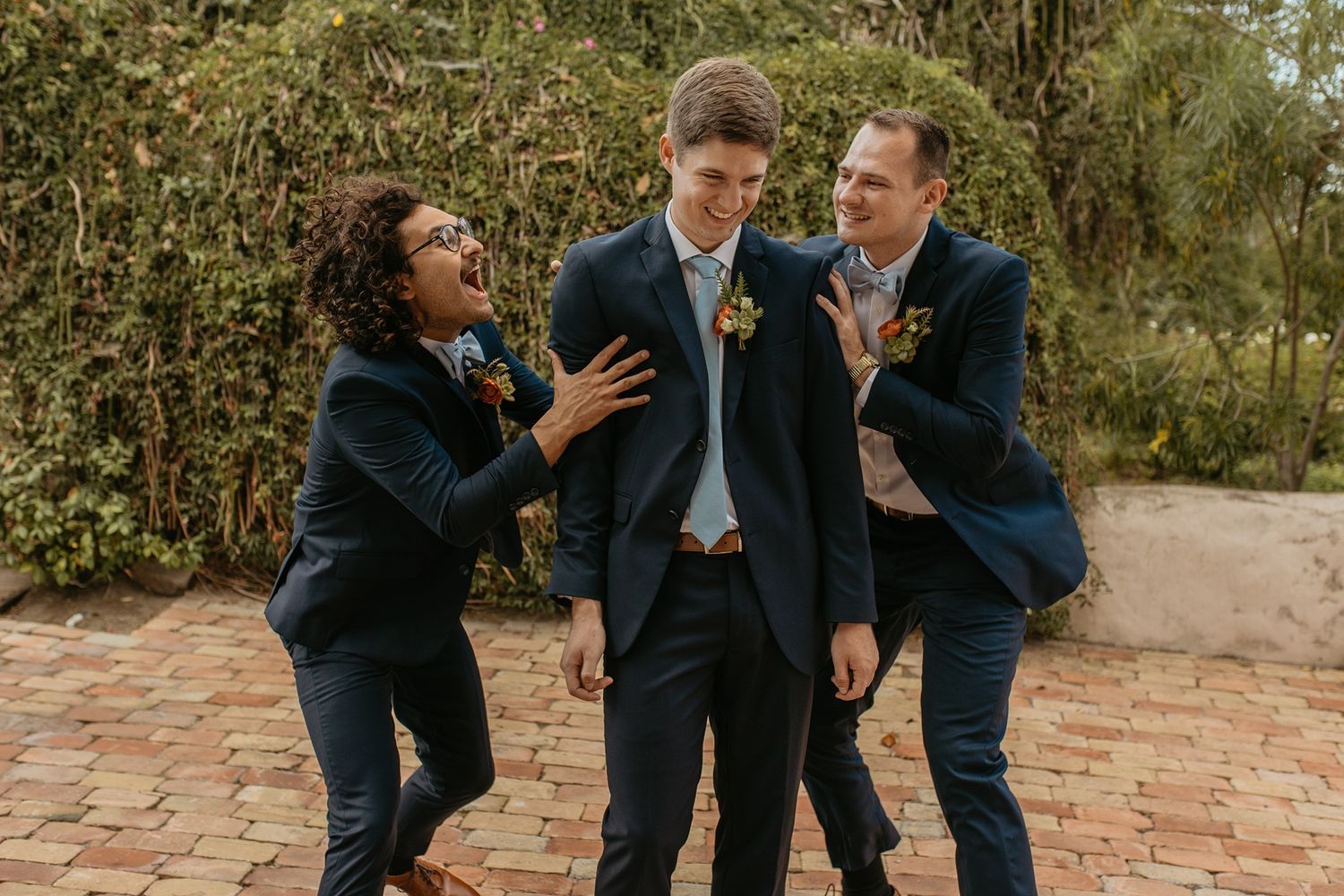 Three people in navy suits with boutonnieres laugh together on a brick patio at a wedding celebration at Rancho Buena Vista Adobe.