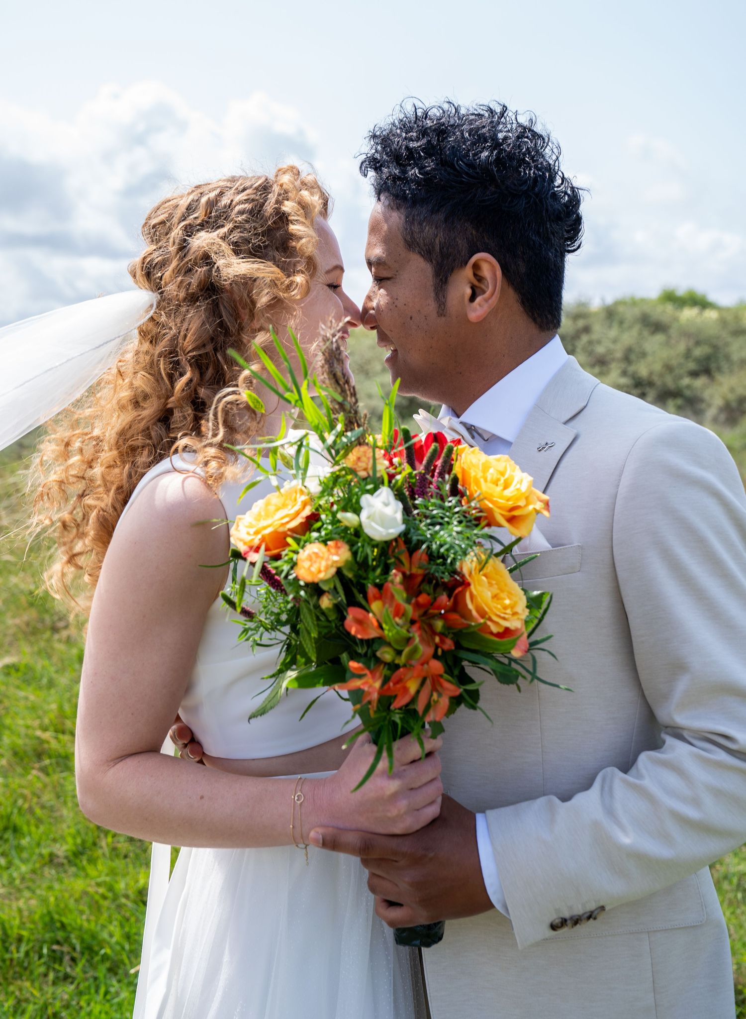 Bruid en bruidegom delen een intiem moment in de buitenlucht, terwijl ze een kleurrijk boeket oranje en gele rozen vasthouden.