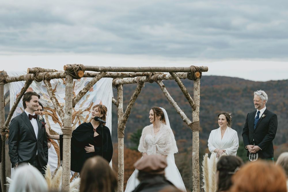 A rustic outdoor wedding ceremony under a wooden arch with mountains in the background.