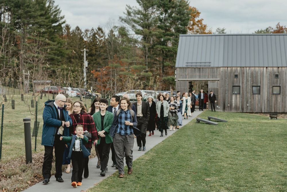 Wedding guests gather outdoors on a grassy path surrounded by autumn trees and rustic buildings.