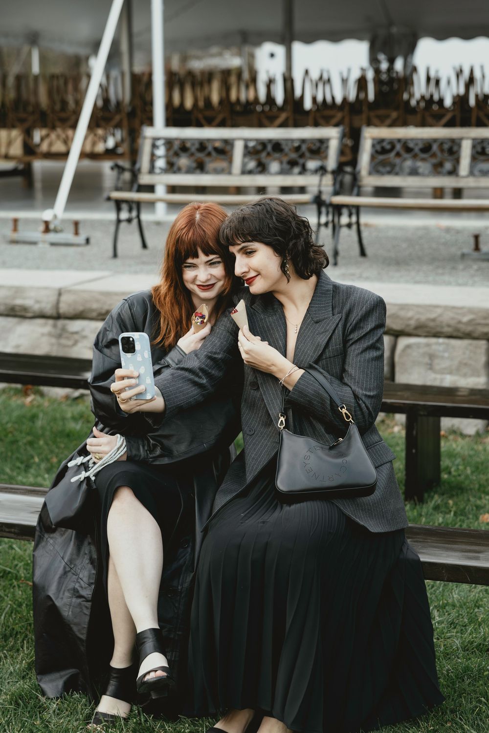 Two friends in dark outfits share a laugh while looking at a phone together on a park bench.