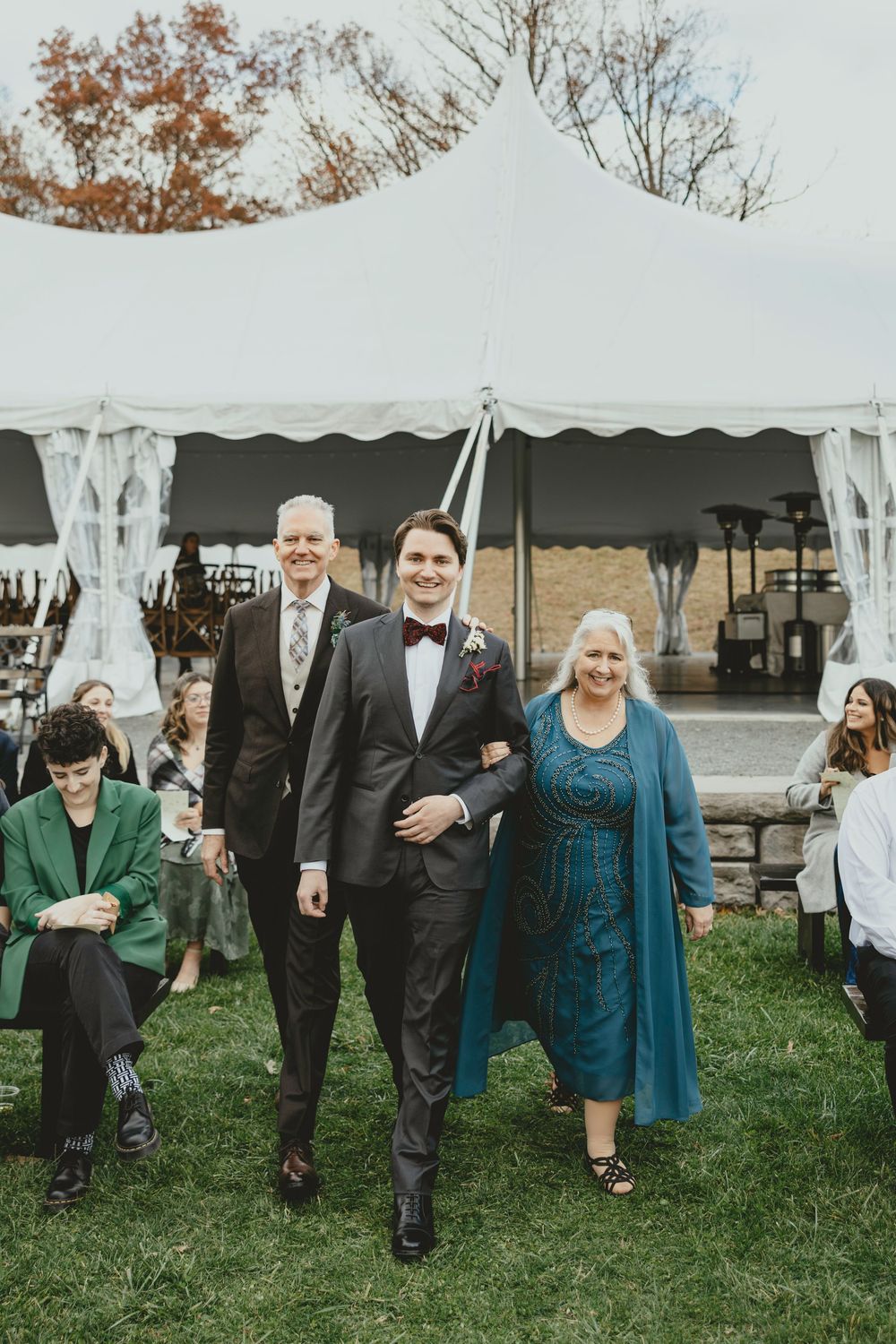Wedding party walking across lawn under elegant white tent during autumn outdoor celebration.
