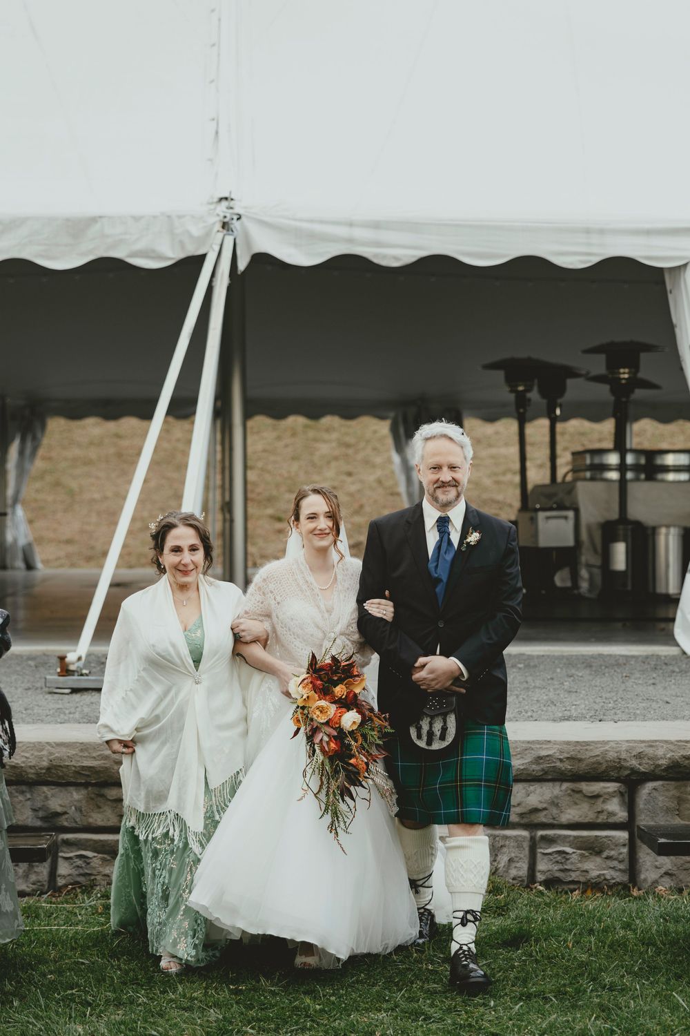 A wedding party gathers under a white tent canopy on a grassy lawn with rustic stone walls in the background.