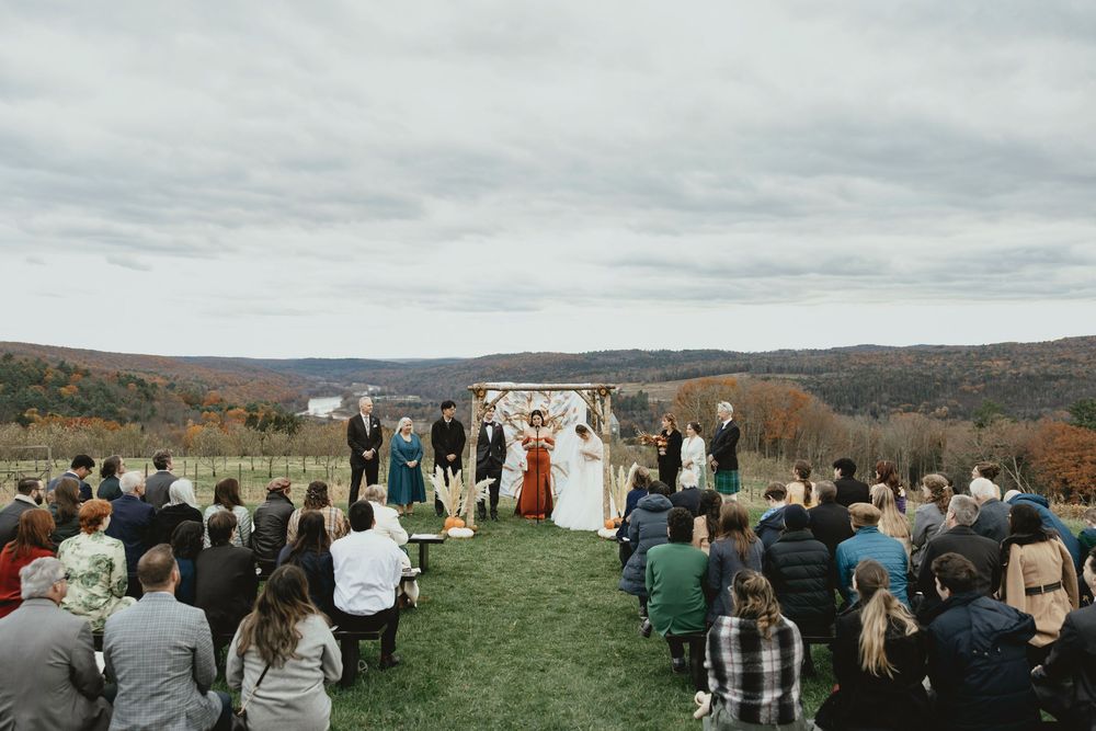 Wedding guests gather outdoors on a hilltop overlooking a scenic mountain landscape during an autumn ceremony.