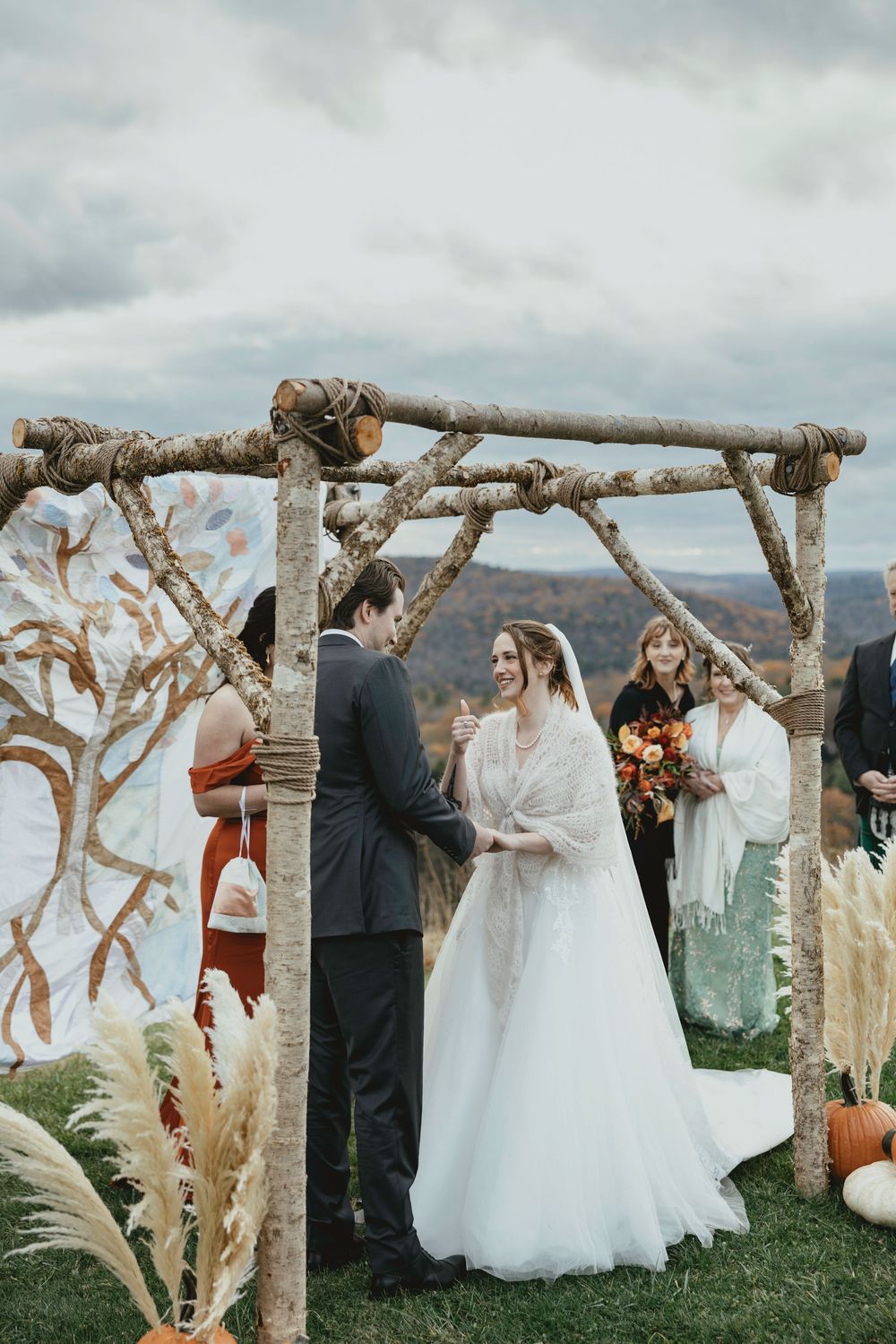 A rustic outdoor wedding ceremony takes place under a natural driftwood arch with mountains in the background.