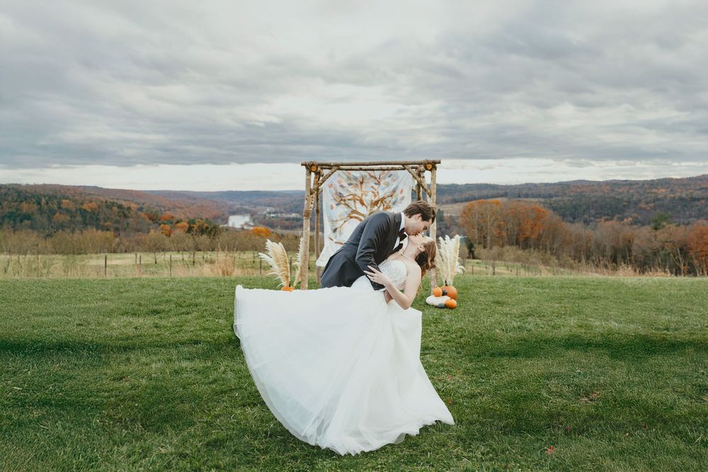 A couple in wedding attire embraces in a scenic autumn field with rolling hills and fall foliage in the background.