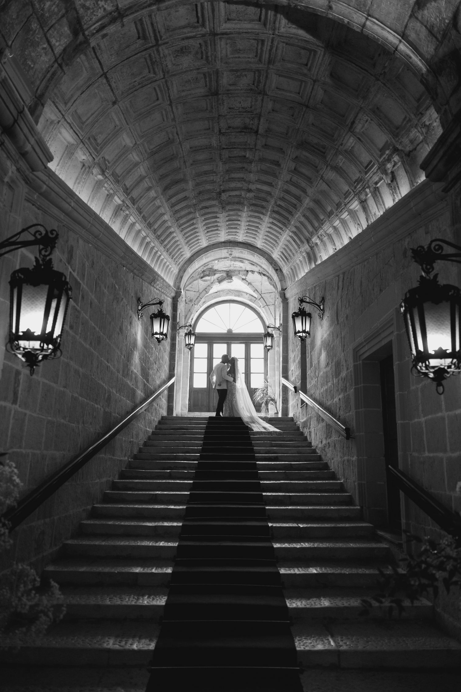 A black and white series of stone stairways with arched ceilings and wall-mounted lanterns leading to bright windows.