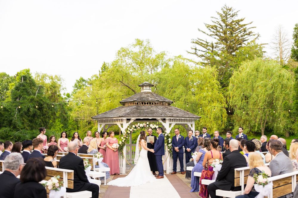 Outdoor wedding ceremony taking place under a wooden gazebo surrounded by lush green trees and guests seated on white chairs.