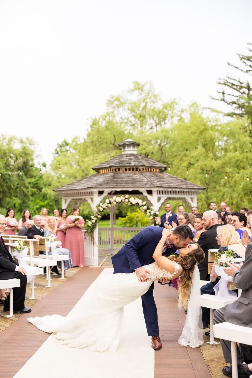 Wedding couple performs romantic dip kiss on outdoor deck with gazebo and guests in background during reception.