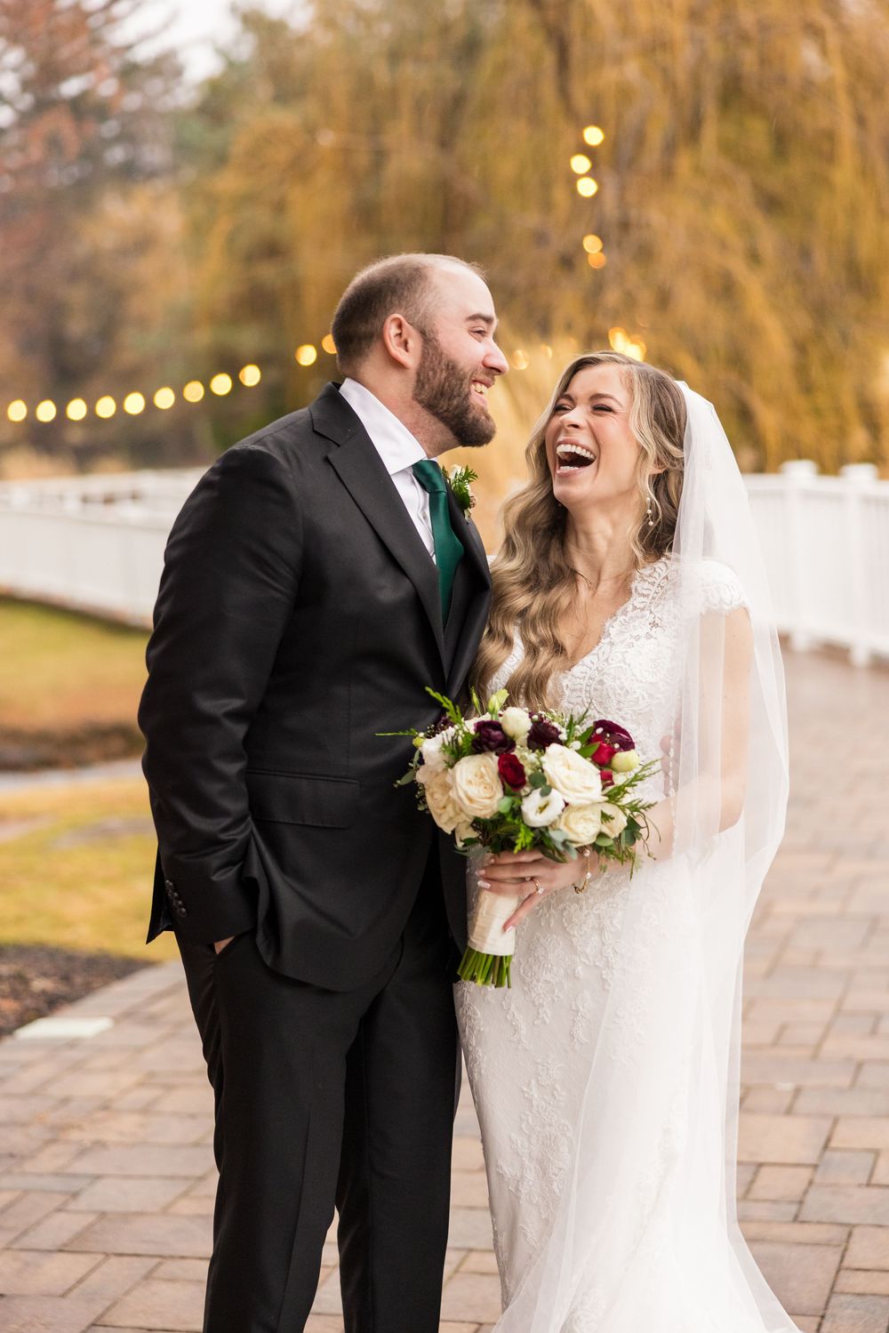 A joyful wedding couple laughs together on a stone patio with autumn trees and string lights in the background.