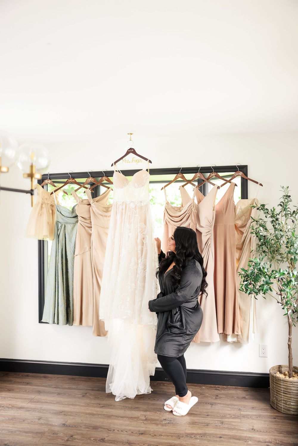 Neutral-toned bridesmaid dresses and a white wedding gown displayed on hangers against a white wall in a bridal shop.