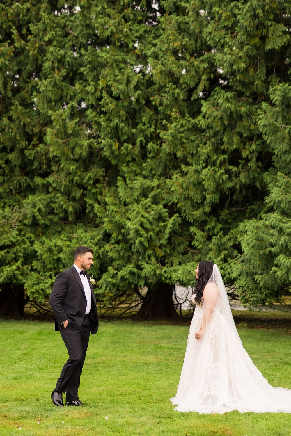 A wedding couple poses for photos on a green lawn with lush trees in the background.