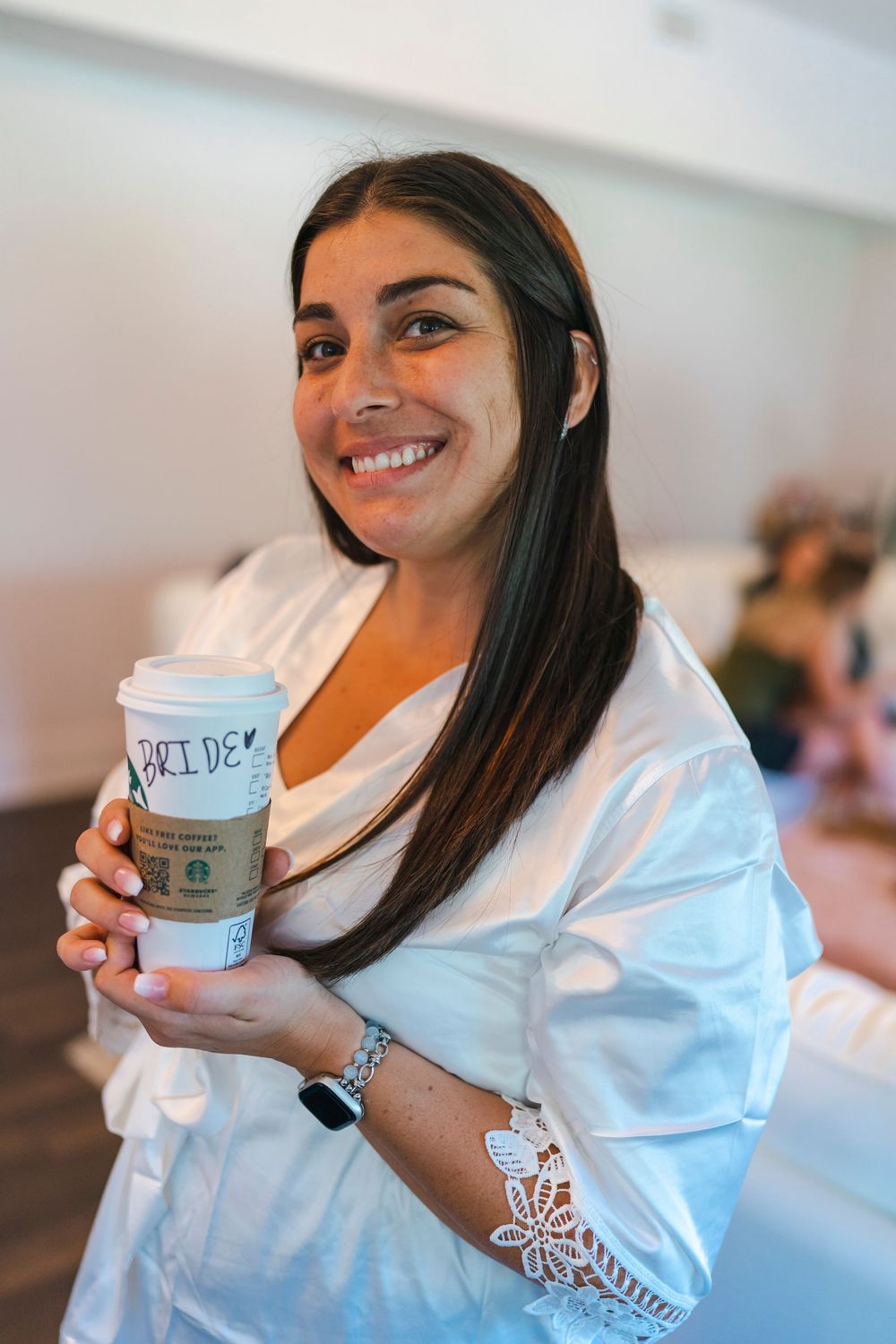 A woman in a white top holds a Starbucks coffee cup while smiling at the camera.