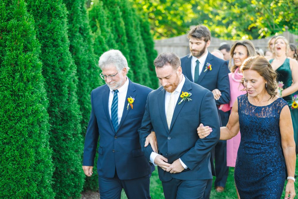 Wedding party members walk through a garden path lined with tall green cypress trees on a sunny day.
