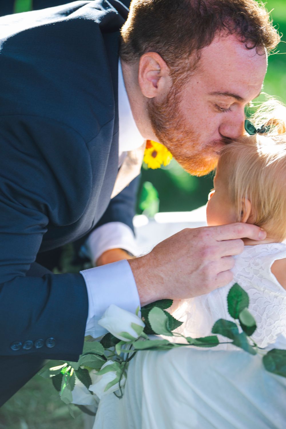A couple shares a romantic kiss in formal wedding attire outdoors with green foliage in the background.