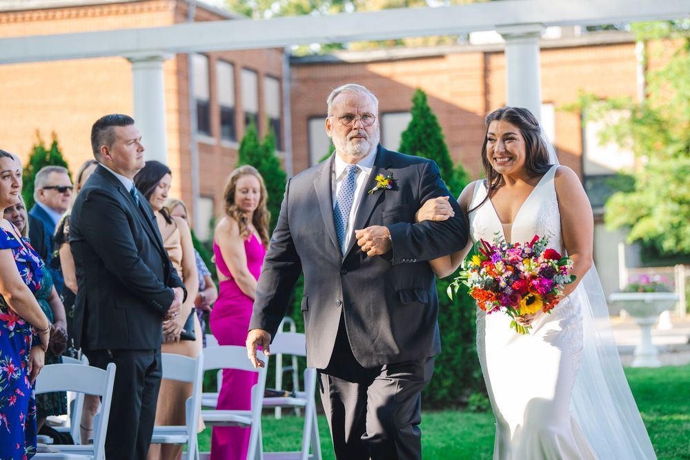A joyful wedding ceremony processional takes place outdoors with colorful floral bouquets and white chairs.