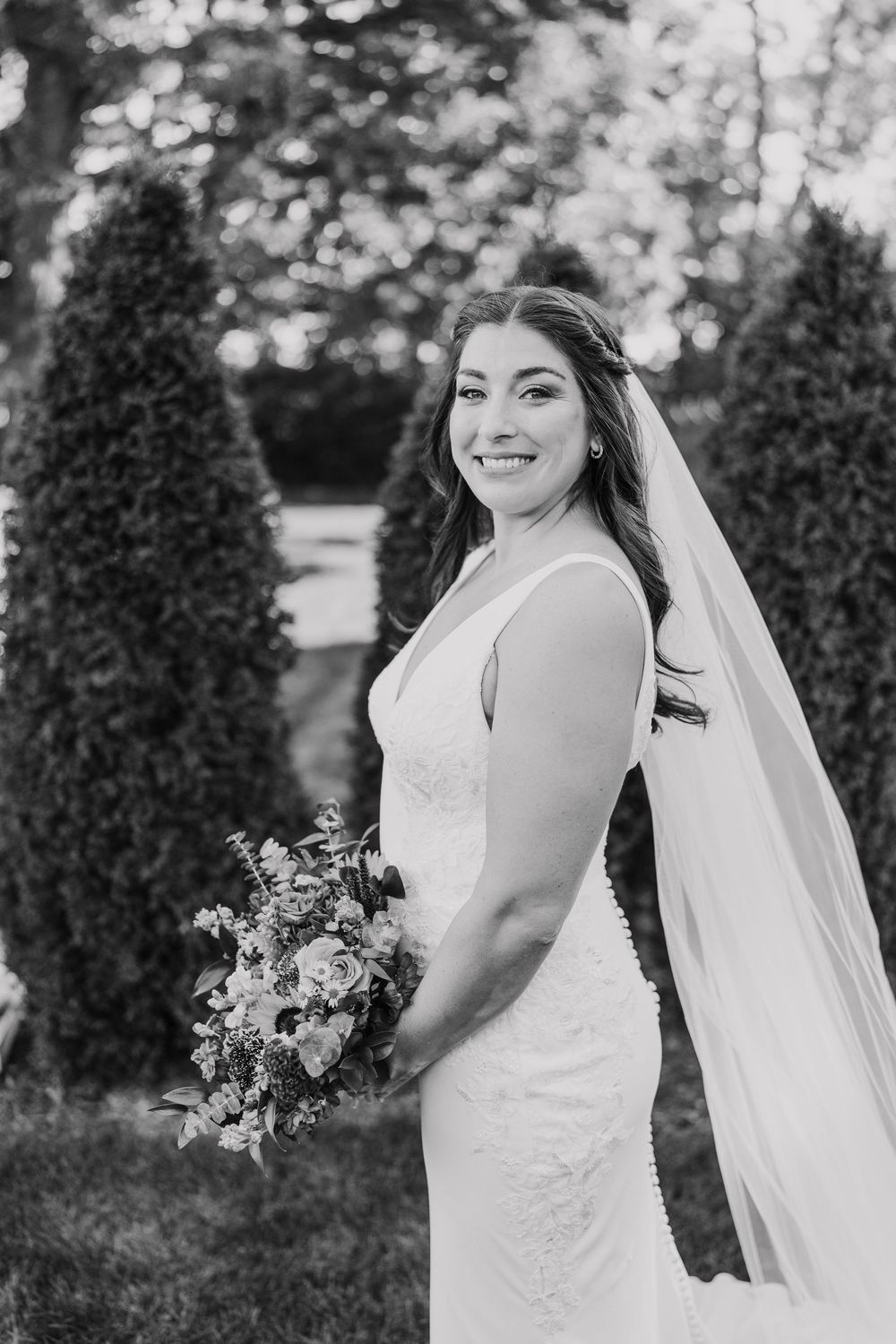 A radiant bride in a fitted white dress poses with her bouquet against a backdrop of evergreen trees.