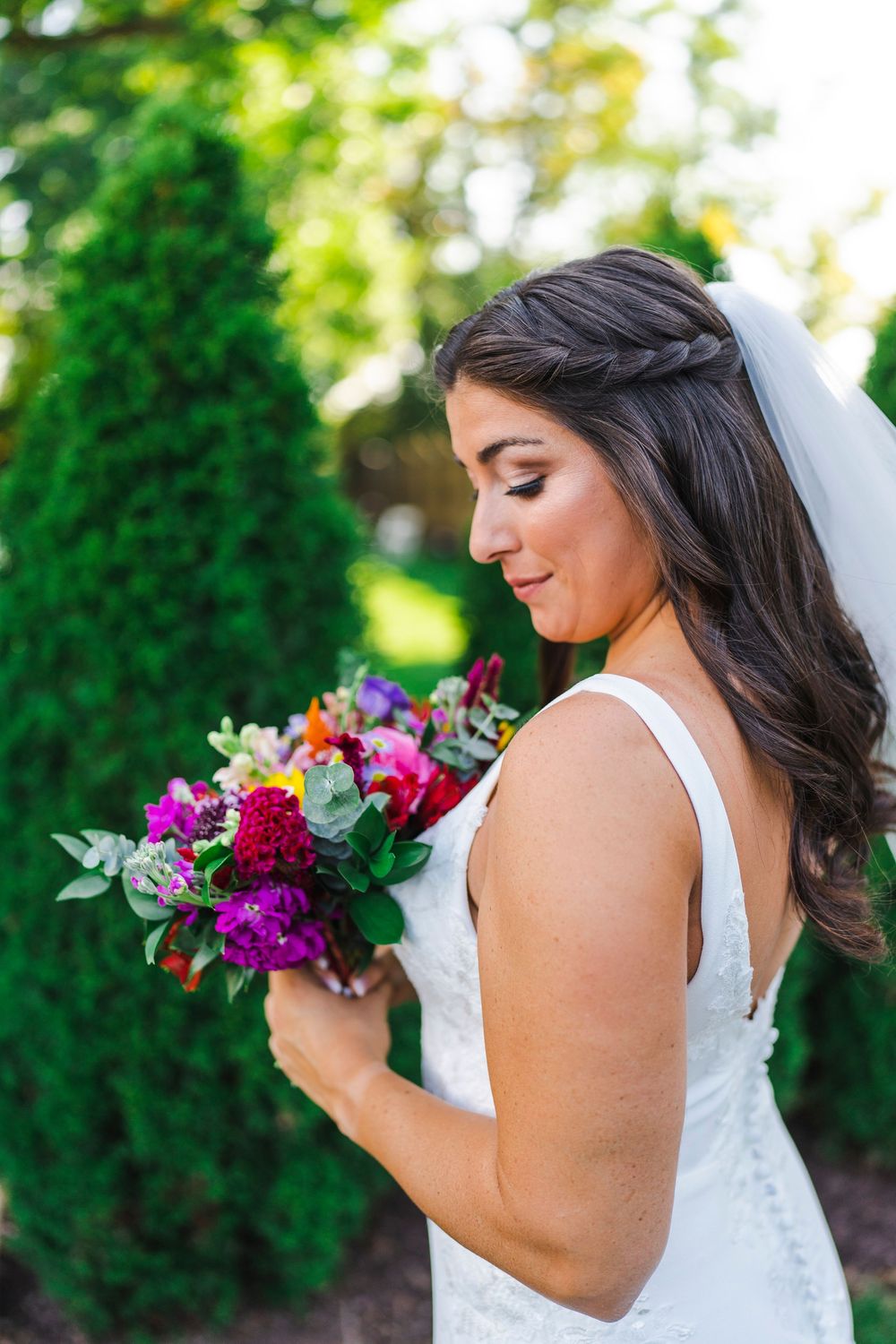 A bride in a white dress holds a vibrant purple and pink wedding bouquet in a garden setting.