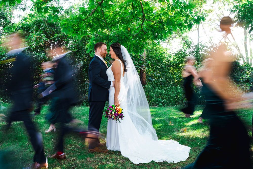 A wedding couple poses while guests create motion blur walking past in a garden setting.