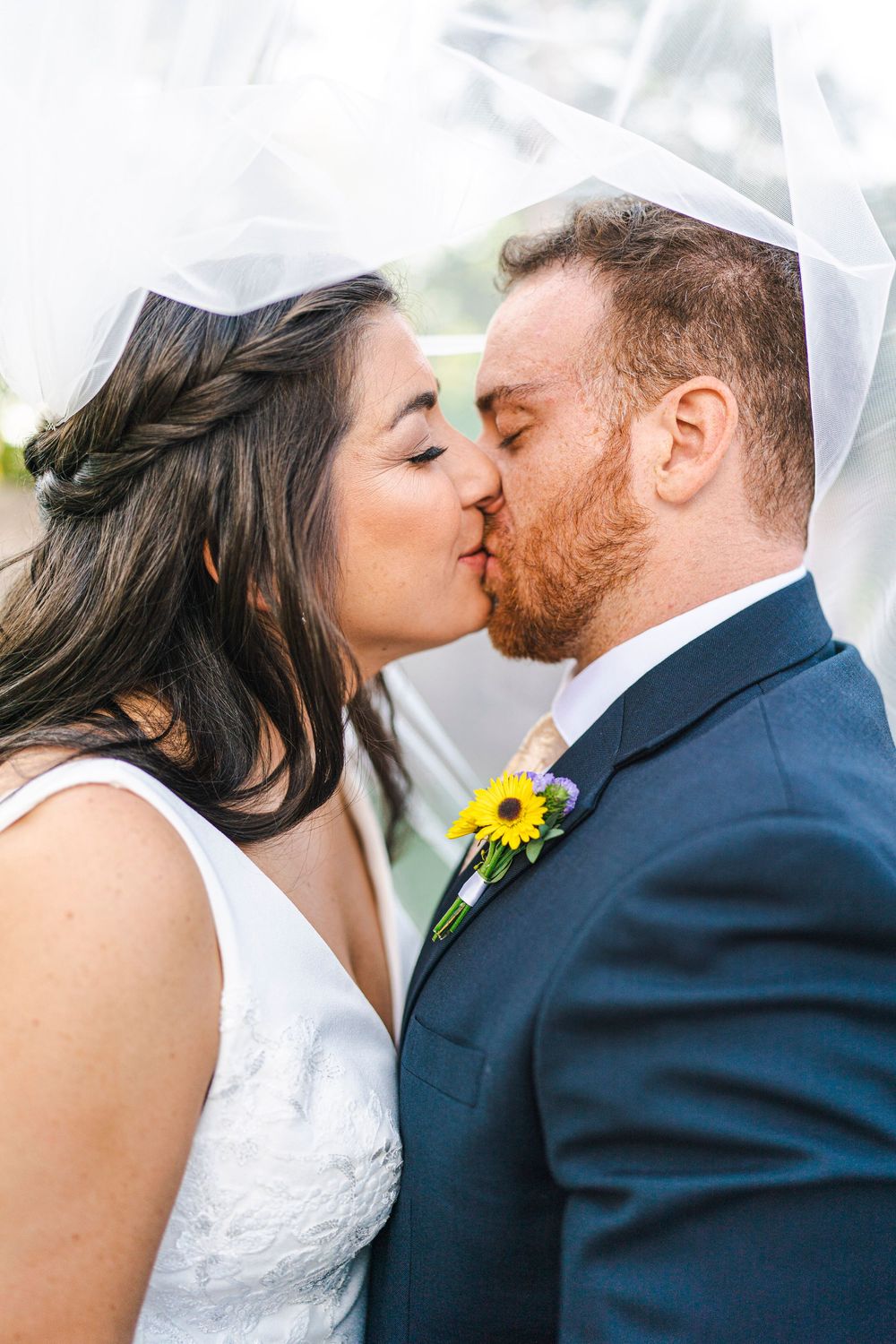 A bride and groom share a romantic kiss under a veil with a sunflower boutonniere.