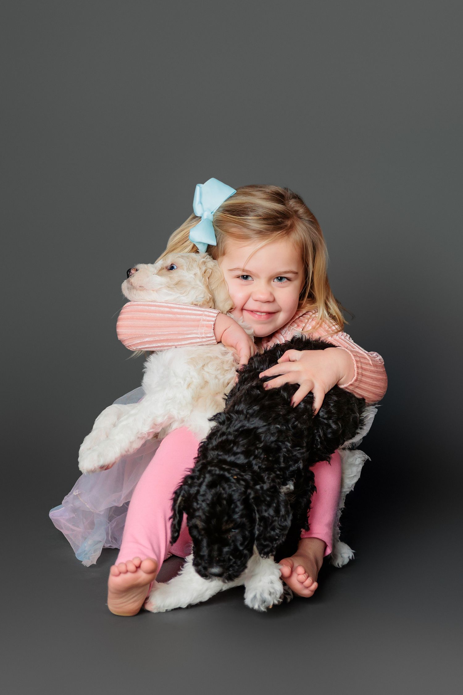 A young child in pink hugs two fluffy dogs - one black and one white - against a dark studio background.