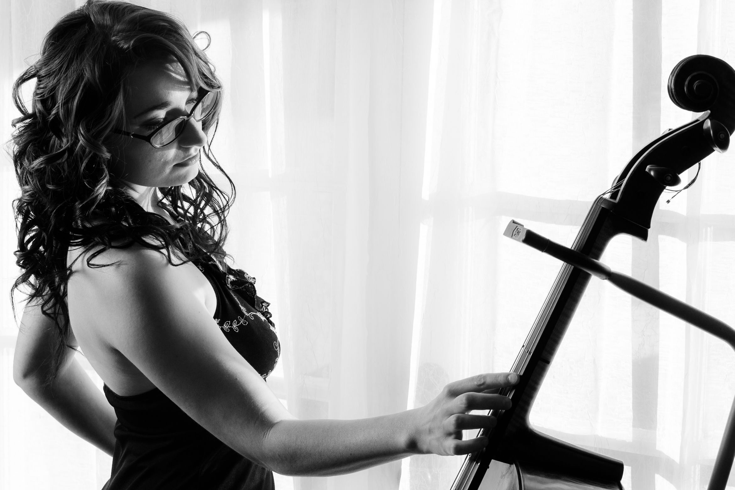 Silhouetted musician in black dress plays cello by window in dramatic black and white photograph.