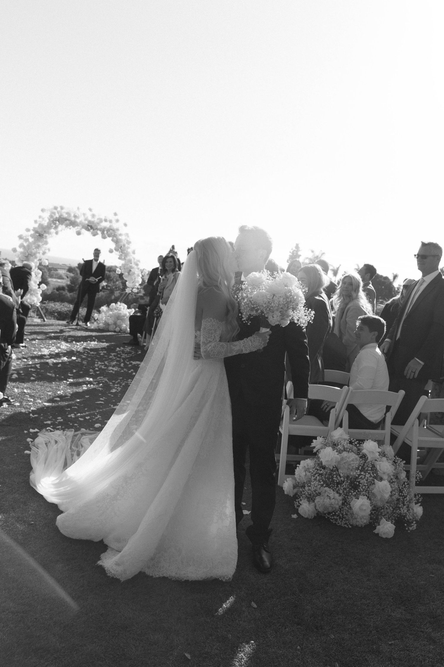 A bride in a flowing lace wedding dress stands on a crosswalk in black and white.