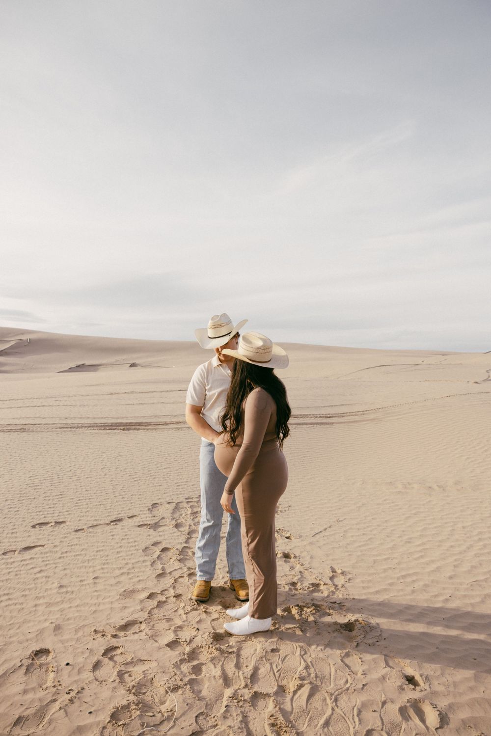 Couple in casual attire embracing on sandy dunes against cloudy sky.
