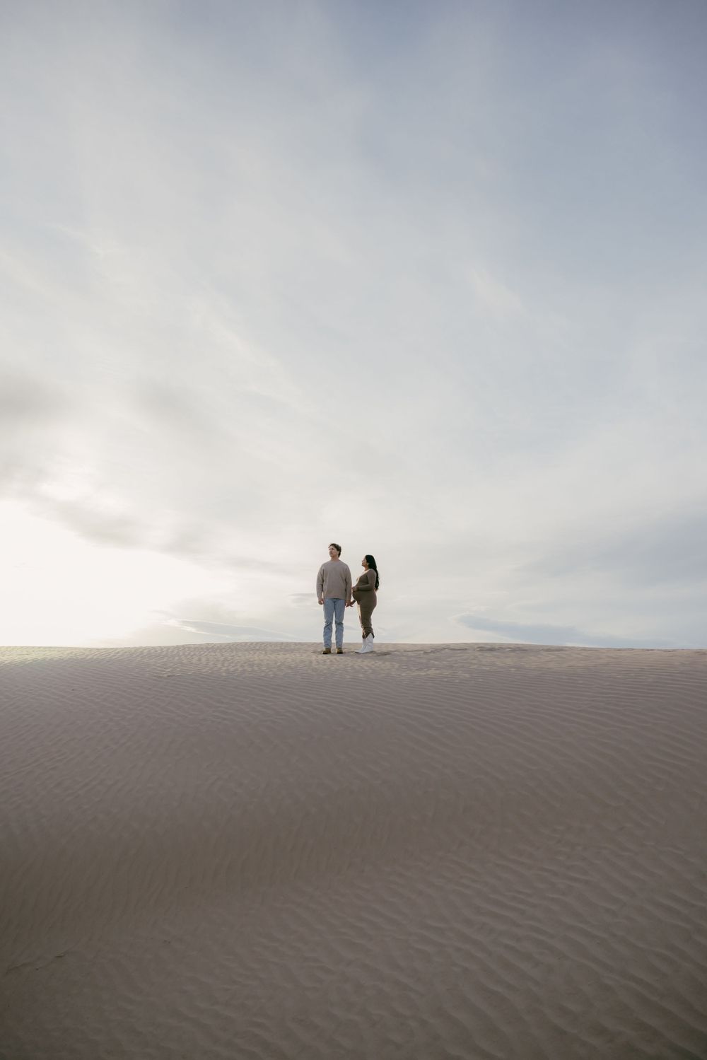 Two silhouettes standing atop desert sand dune against cloudy sky.