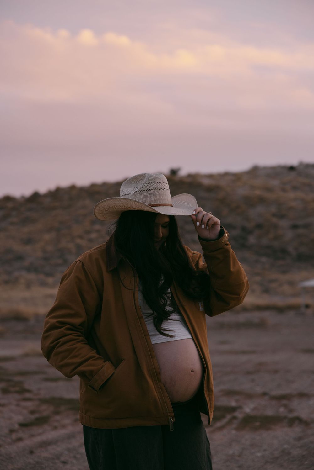 Maternity photo at sunset with cowboy hat and brown jacket in desert setting.