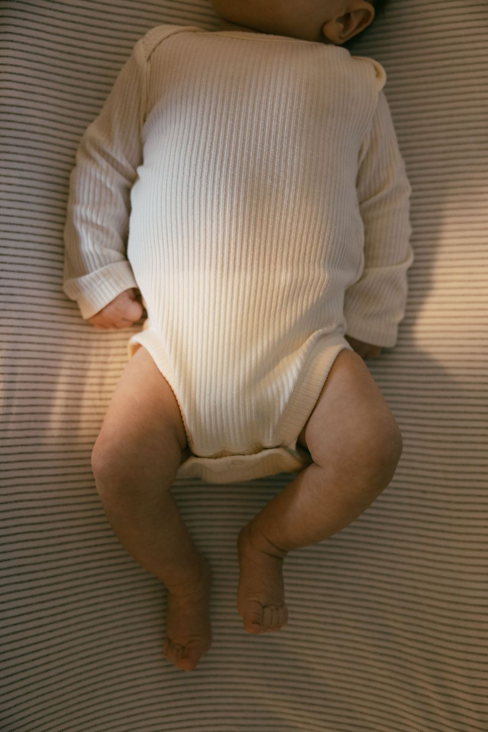 An infant wearing a white ribbed onesie lies on striped bedding.