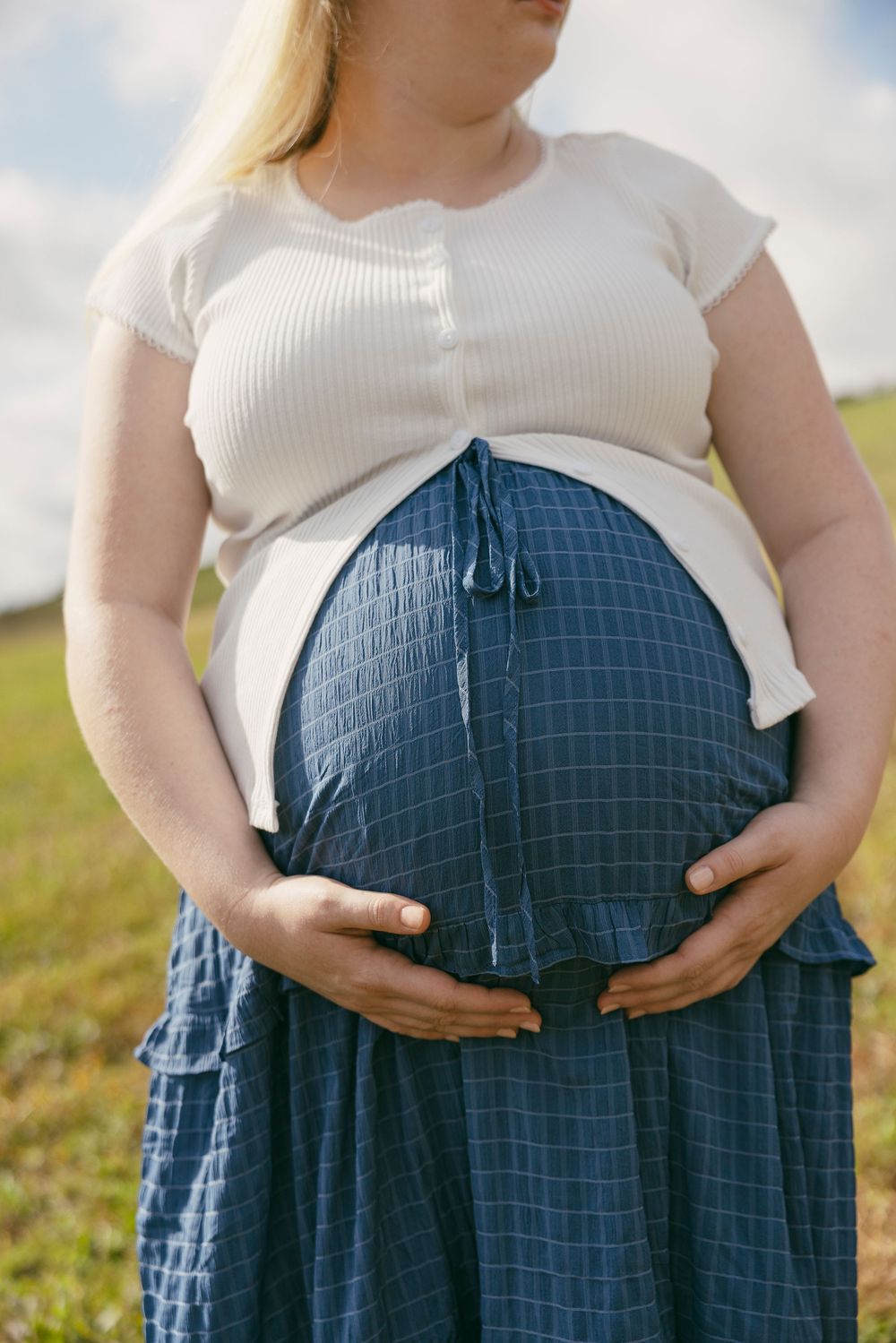 Pregnant woman in white ribbed top and blue plaid skirt cradling her baby bump outdoors in a green field.