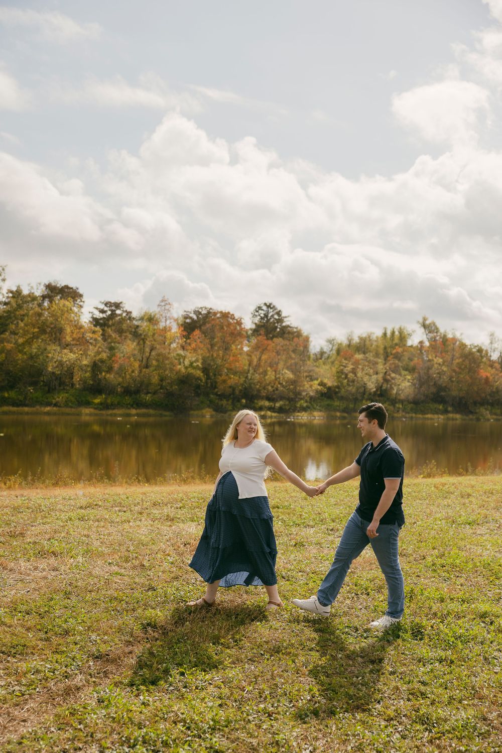 Couple holding hands and walking on grass near a lake with autumn trees and cloudy sky in background.
