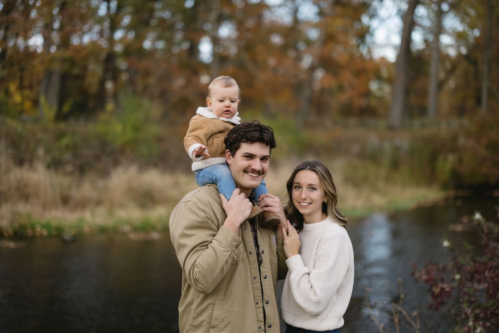 A family poses together by a pond surrounded by autumn trees and fall colors.