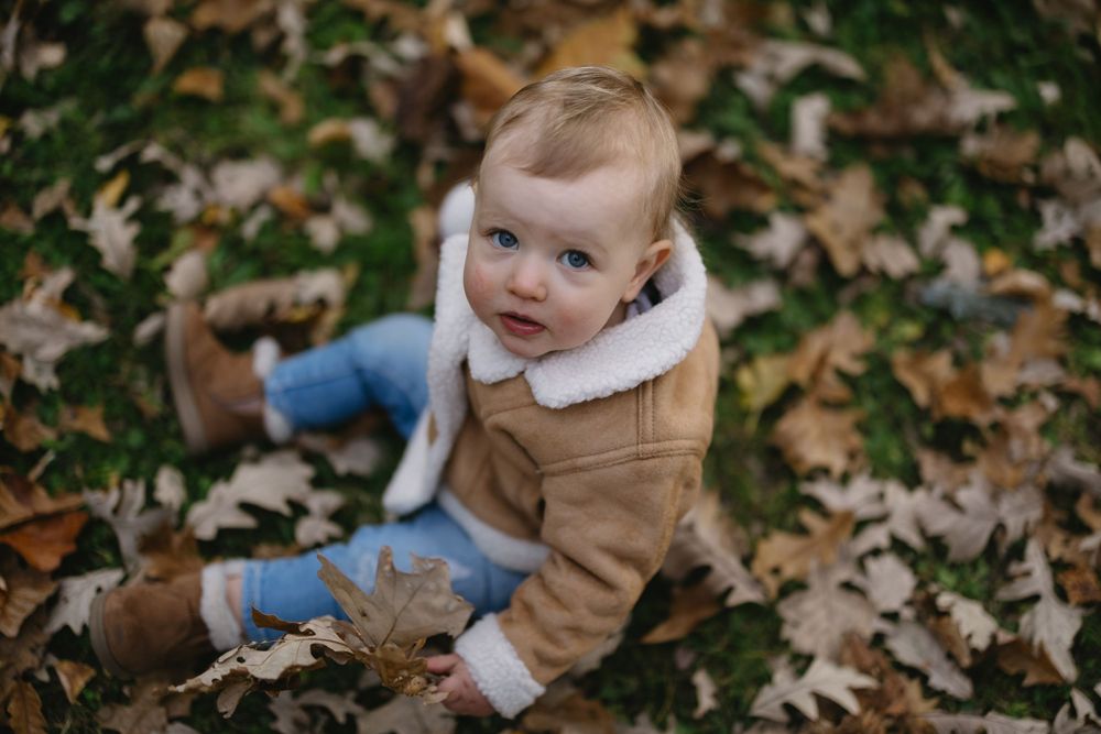 A baby in a brown jacket sits among fallen autumn leaves looking curiously at the camera.