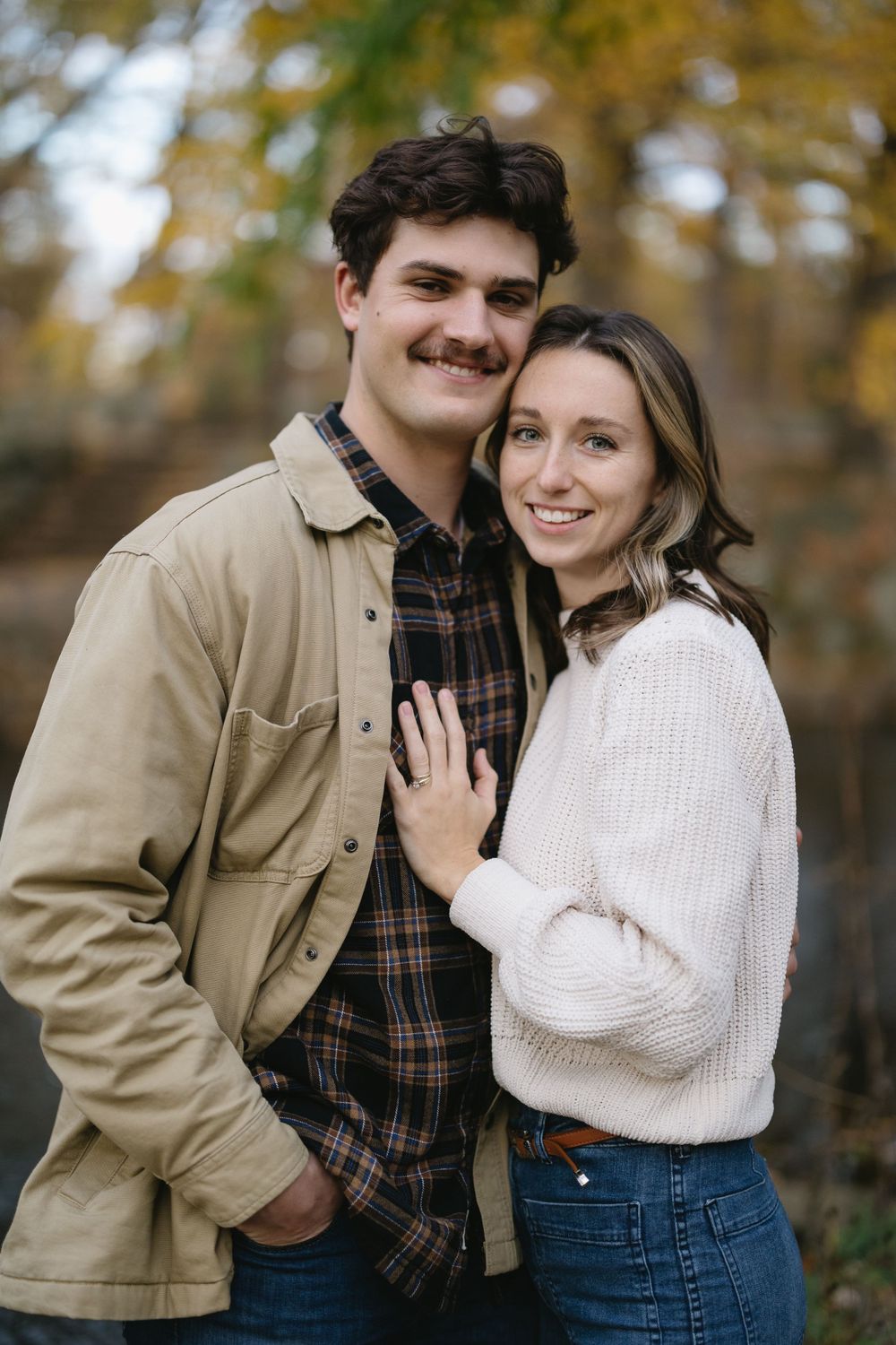 A couple in casual fall attire poses together against an autumn forest backdrop.