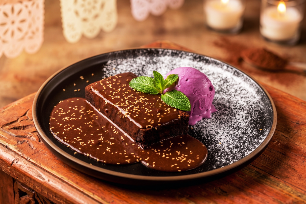 Chocolate brownie slice topped with powdered sugar and mint, served with purple berry ice cream on a dark plate.