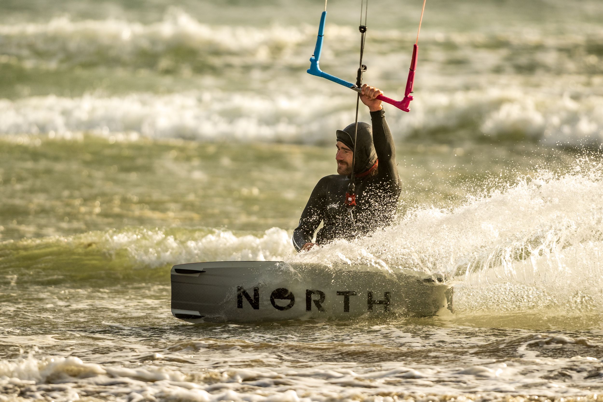 A kitesurfer rides the waves while performing aerial tricks at sunset with splashing water in the background.
