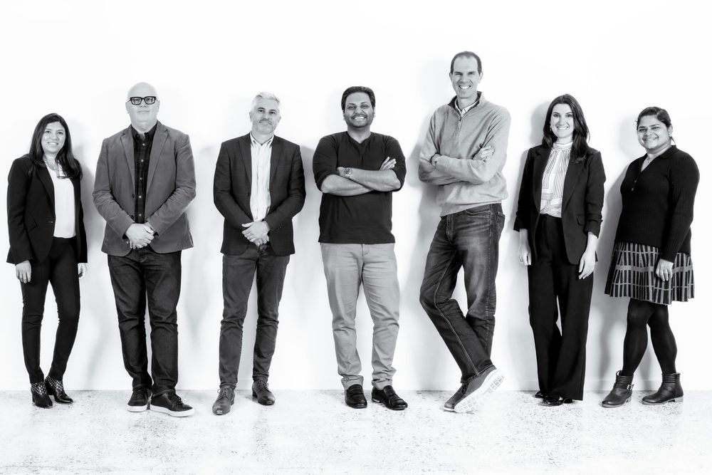Black and white photo of seven diverse business professionals standing together in a modern studio setting.