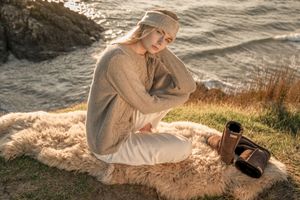 Woman in beige outfit relaxes on sandy beach at sunset with water in background.