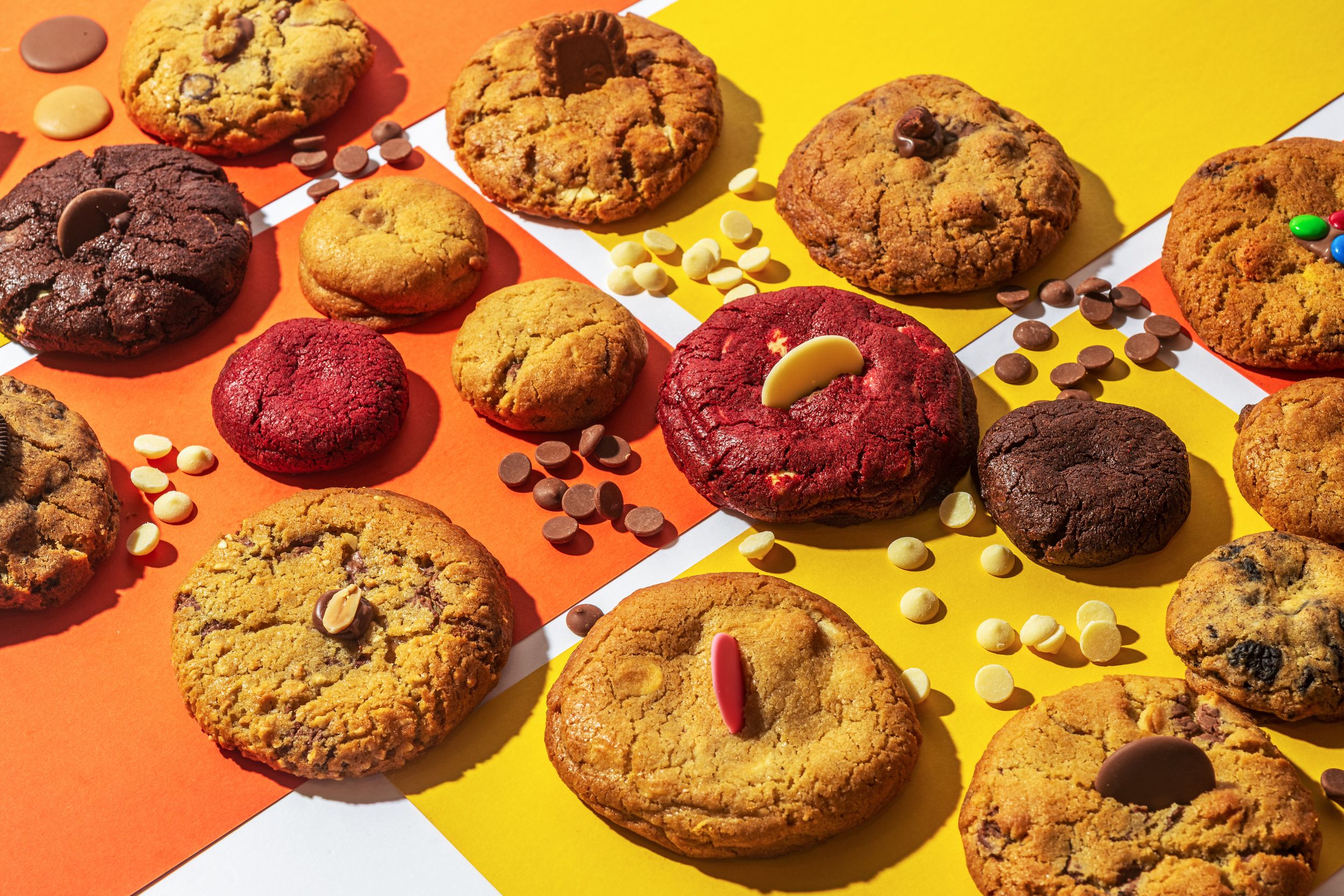 Assorted homemade cookies with chocolate chips, nuts, and red velvet varieties arranged on orange and yellow background.