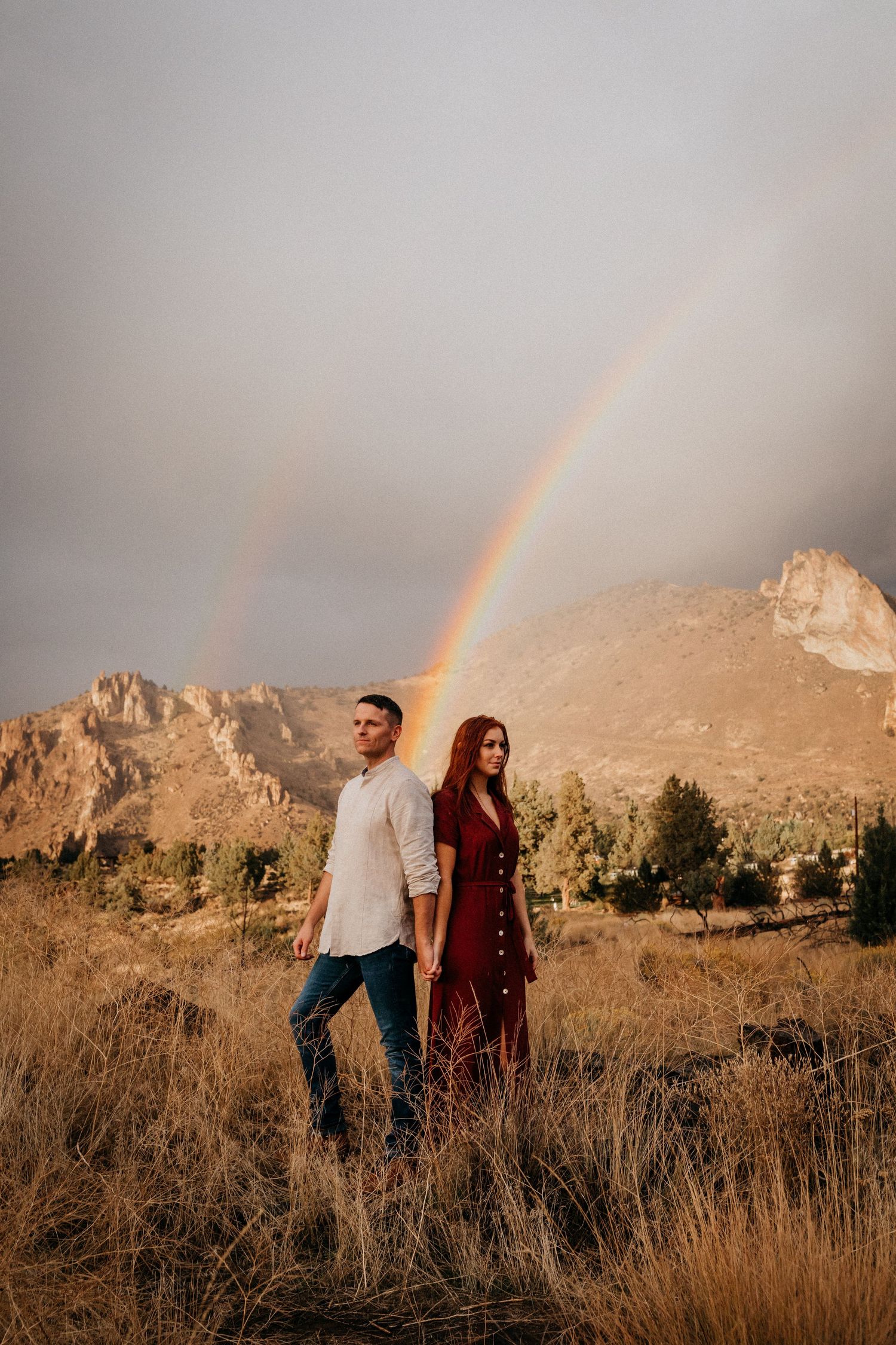 A couple stands together in a scenic mountain landscape with a double rainbow arcing overhead in golden evening light.