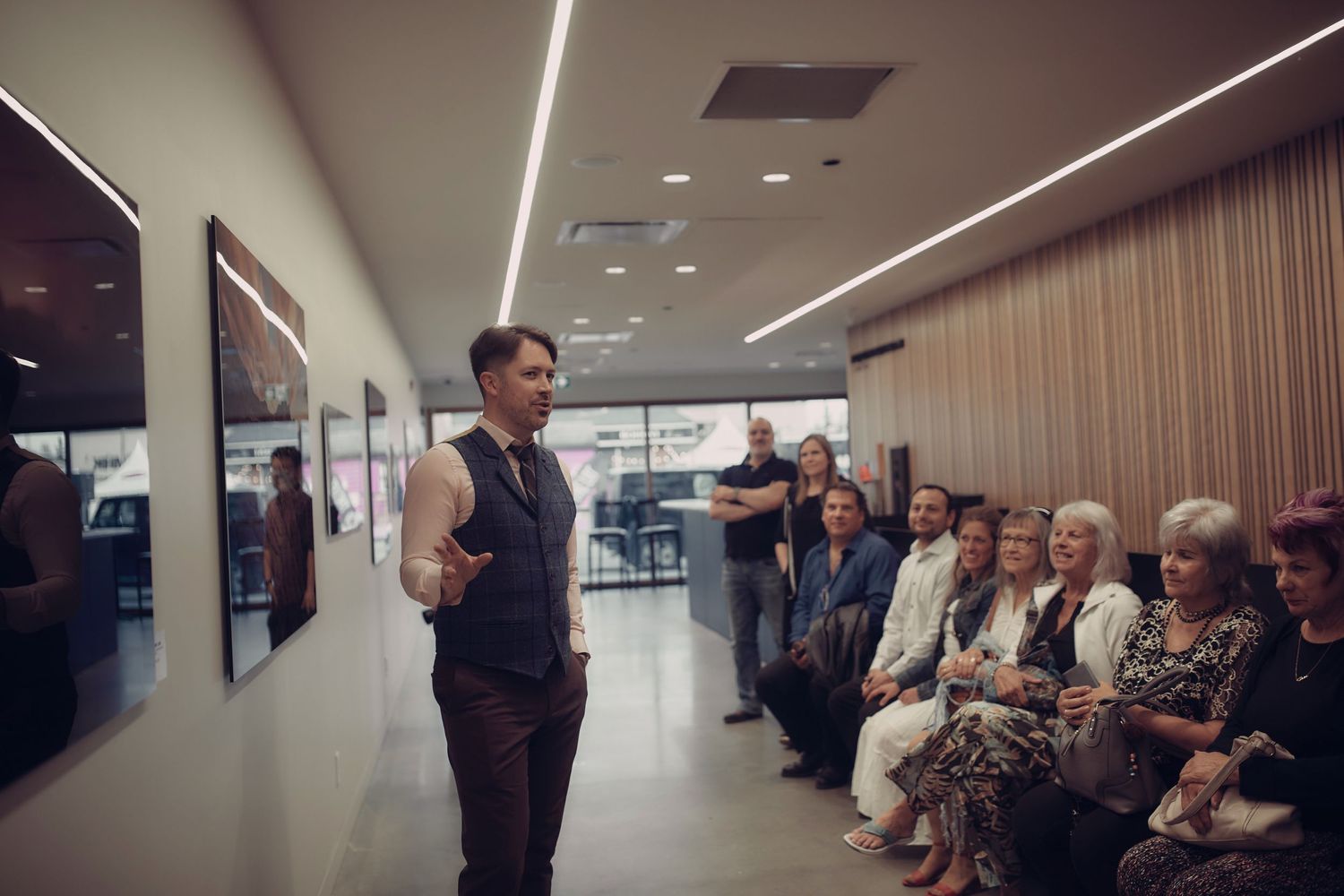 A speaker, Evan Will, in a dark vest addresses an audience seated along a wall in a modern hallway with recessed lighting for his gallery