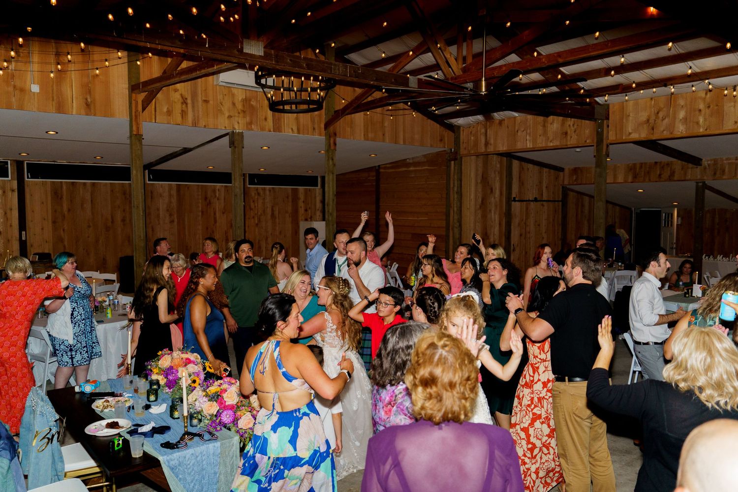 Wedding reception guests dancing in rustic wooden barn venue with string lights.