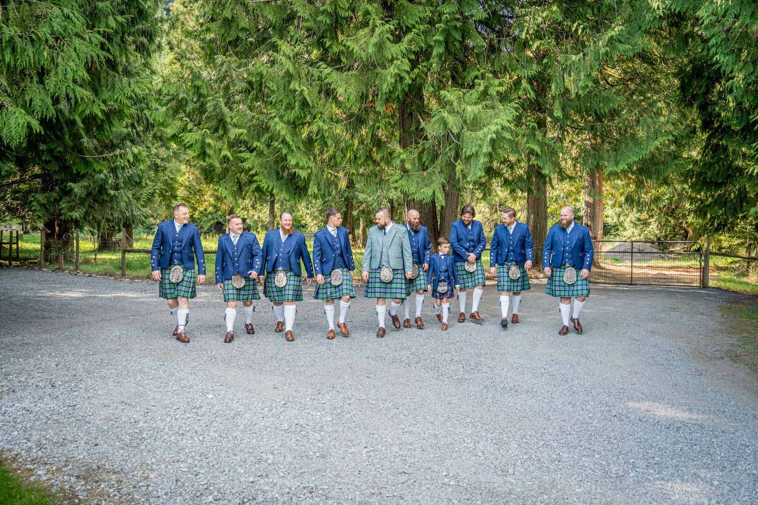 Groomsmen in blue suits and kilts walk together on gravel path.