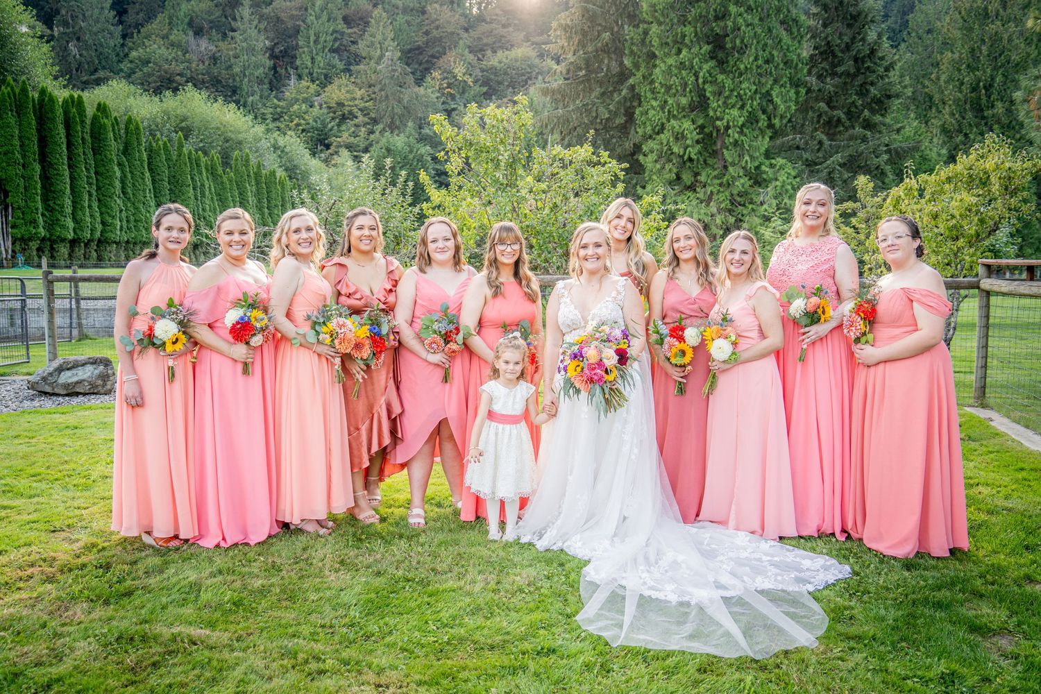 Wedding party in coral pink dresses and white gown pose in front of evergreen trees.