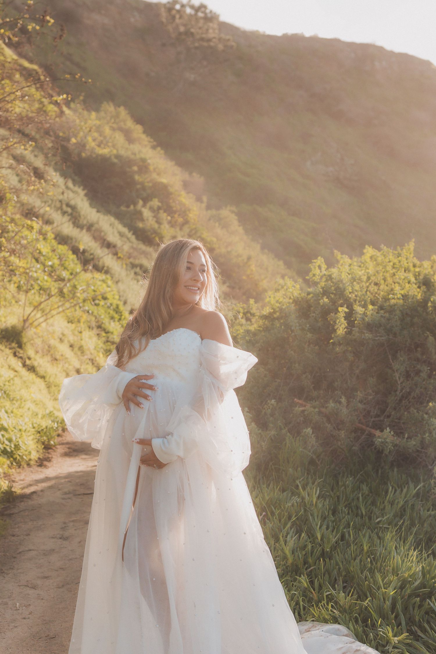 Woman in flowing white dress poses on mountain trail during golden hour.