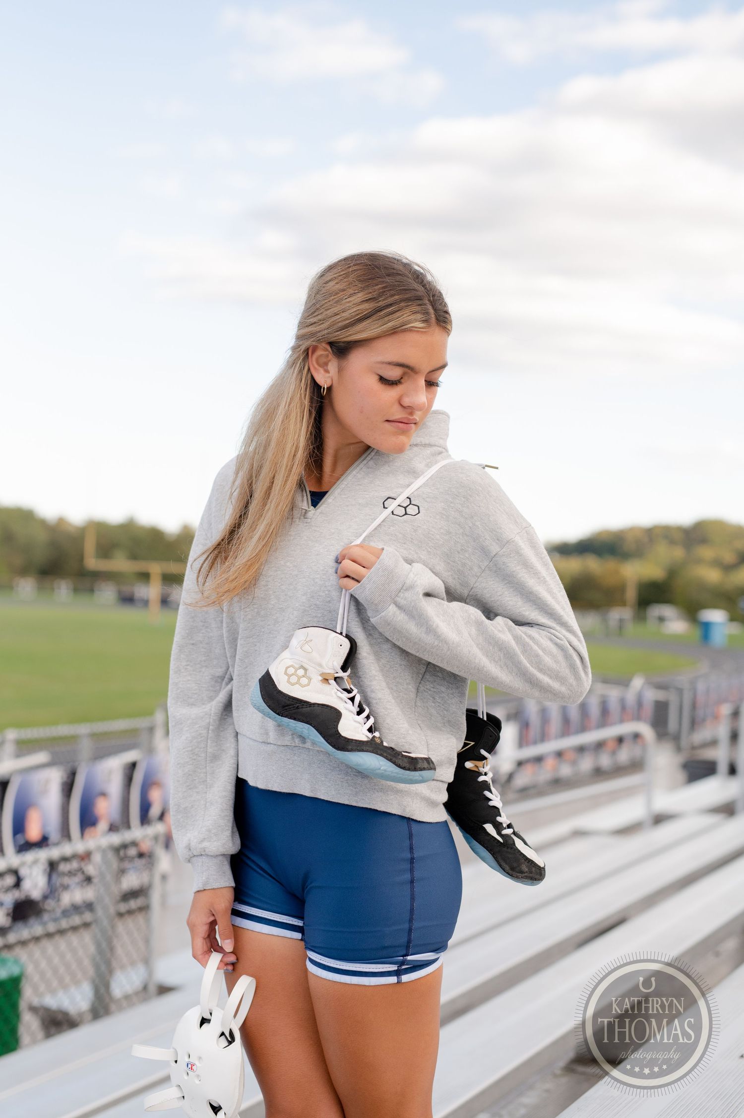 Athlete in grey sweatshirt carries ice skates while standing on stadium bleachers.