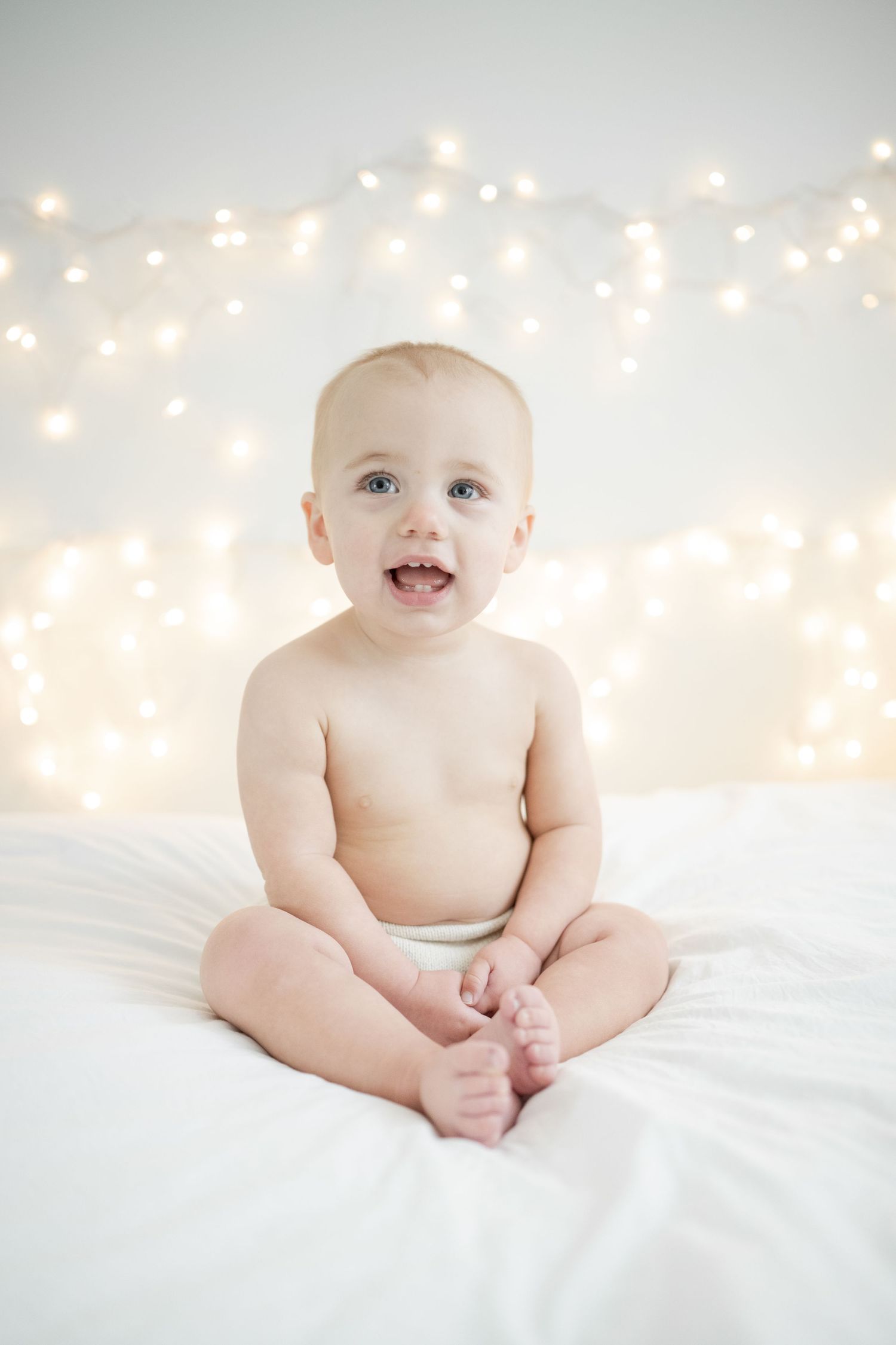 baby girl smiles while sitting on white bedding with twinkling lights behind her - one year milestone session